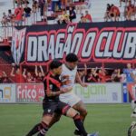 Imagem dois jogadores disputando a balo, ao fundo torcedores no estádio