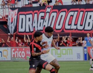 Imagem dois jogadores disputando a balo, ao fundo torcedores no estádio