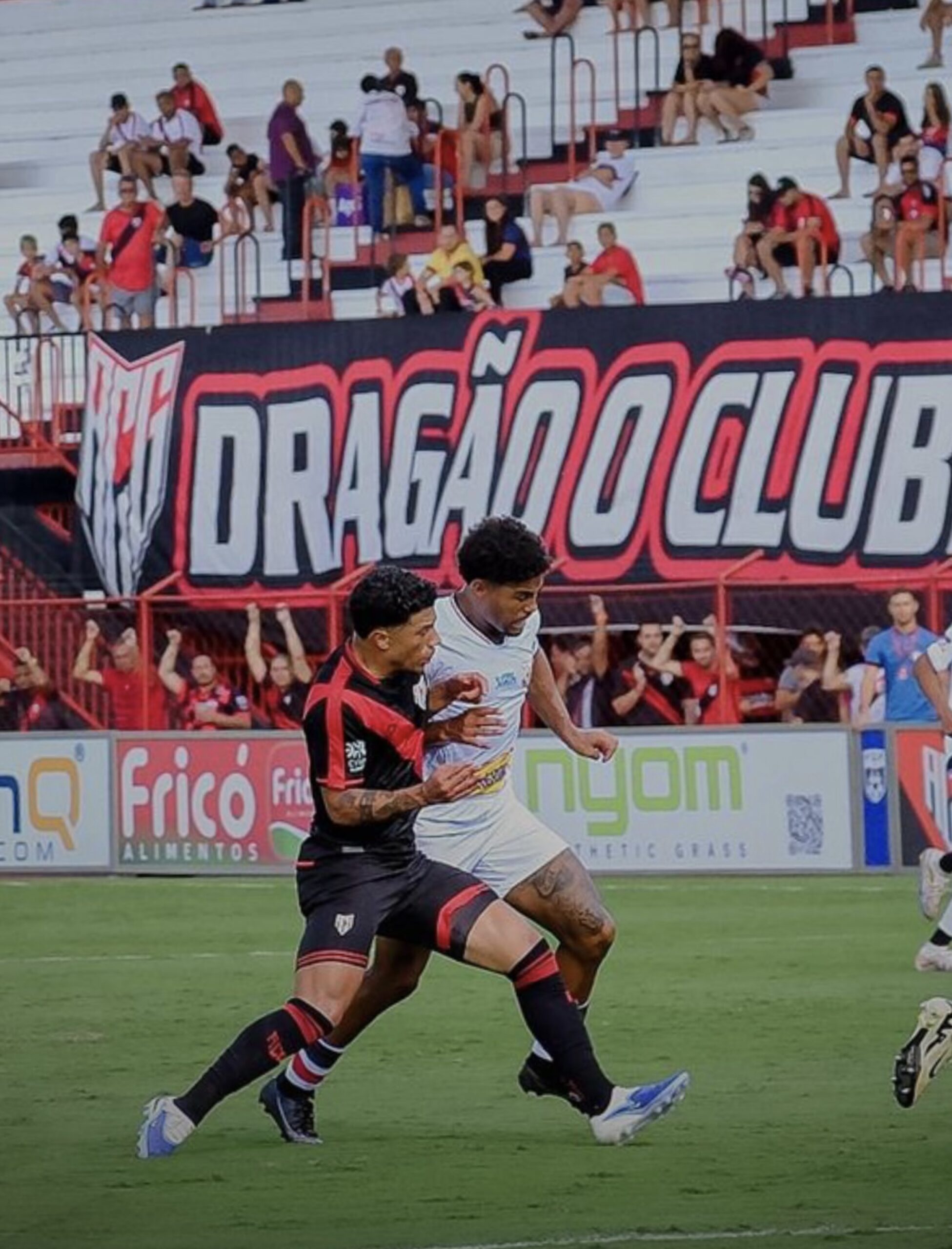 Imagem dois jogadores disputando a balo, ao fundo torcedores no estádio