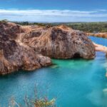 imagem retrata uma paisagem natural de grande impacto visual, formada por um lago de águas azul-esverdeadas intensas, encaixado entre paredões rochosos de tons terrosos e acinzentados. As rochas altas e irregulares mostram marcas evidentes de erosão e escavação, criando um cenári