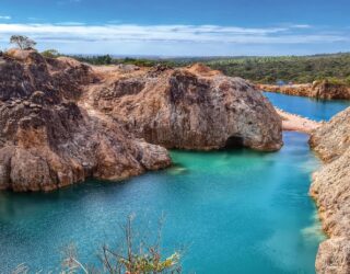 imagem retrata uma paisagem natural de grande impacto visual, formada por um lago de águas azul-esverdeadas intensas, encaixado entre paredões rochosos de tons terrosos e acinzentados. As rochas altas e irregulares mostram marcas evidentes de erosão e escavação, criando um cenári