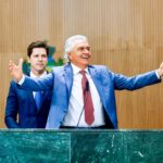 Imagem dois homens de terno um de braços abertos