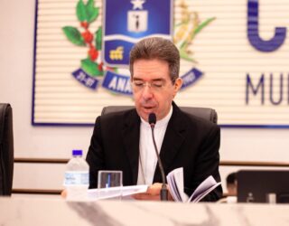 Imagem homem de terno preto sentado falando ao microfone