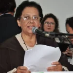 Imagem mulher de óculos segurando papel e falando ao microfone