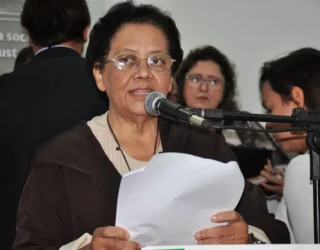 Imagem mulher de óculos segurando papel e falando ao microfone