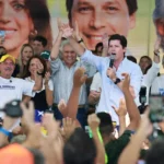 Imagem homem segurando microfone no palco ao lado de várias pessoas