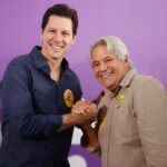 Imagem homem jovem de camisa azul e homem cabelo grisalho de camisa cinza segurando as mãos