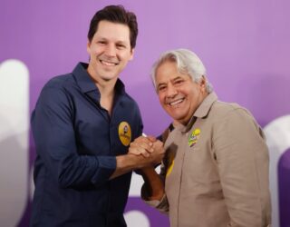 Imagem homem jovem de camisa azul e homem cabelo grisalho de camisa cinza segurando as mãos