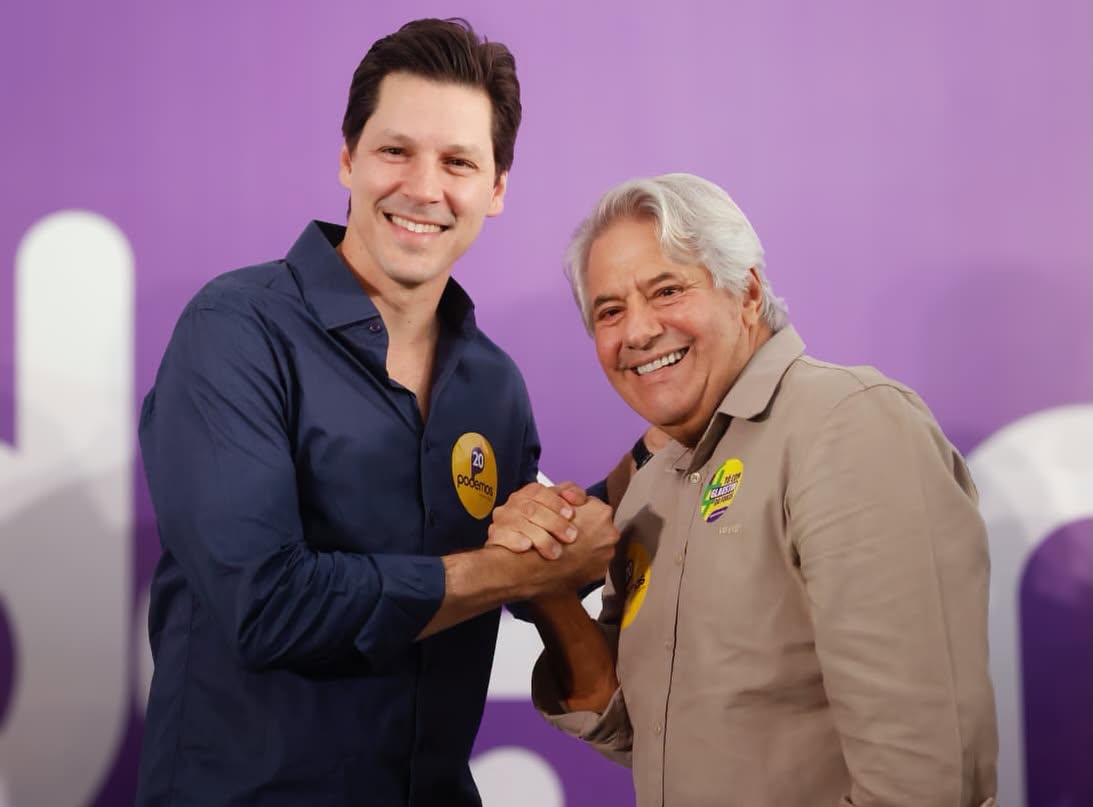 Imagem homem jovem de camisa azul e homem cabelo grisalho de camisa cinza segurando as mãos