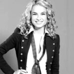Imagem da atriz Lua Blanco em preto e branco. A atriz usa uma camisa branca, com bçazer escuro e gravata, cabelos loiros cacheados e soltos.