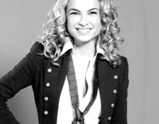 Imagem da atriz Lua Blanco em preto e branco. A atriz usa uma camisa branca, com bçazer escuro e gravata, cabelos loiros cacheados e soltos.