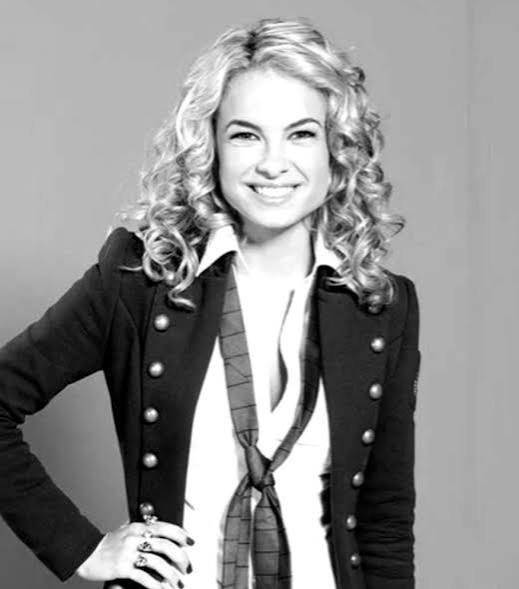 Imagem da atriz Lua Blanco em preto e branco. A atriz usa uma camisa branca, com bçazer escuro e gravata, cabelos loiros cacheados e soltos.