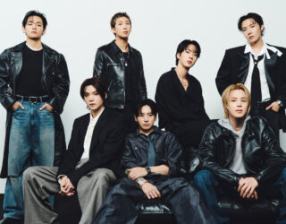 Imagem do BTS, fenômeno mundial. Estão vestindo tons escuros e jeans, três membros sentados e quatro em pé atrás.