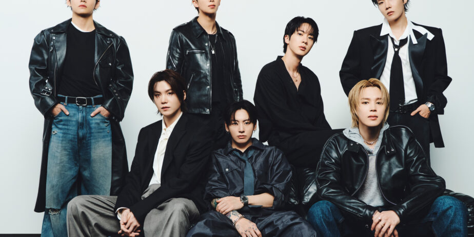 Imagem do BTS, fenômeno mundial. Estão vestindo tons escuros e jeans, três membros sentados e quatro em pé atrás.