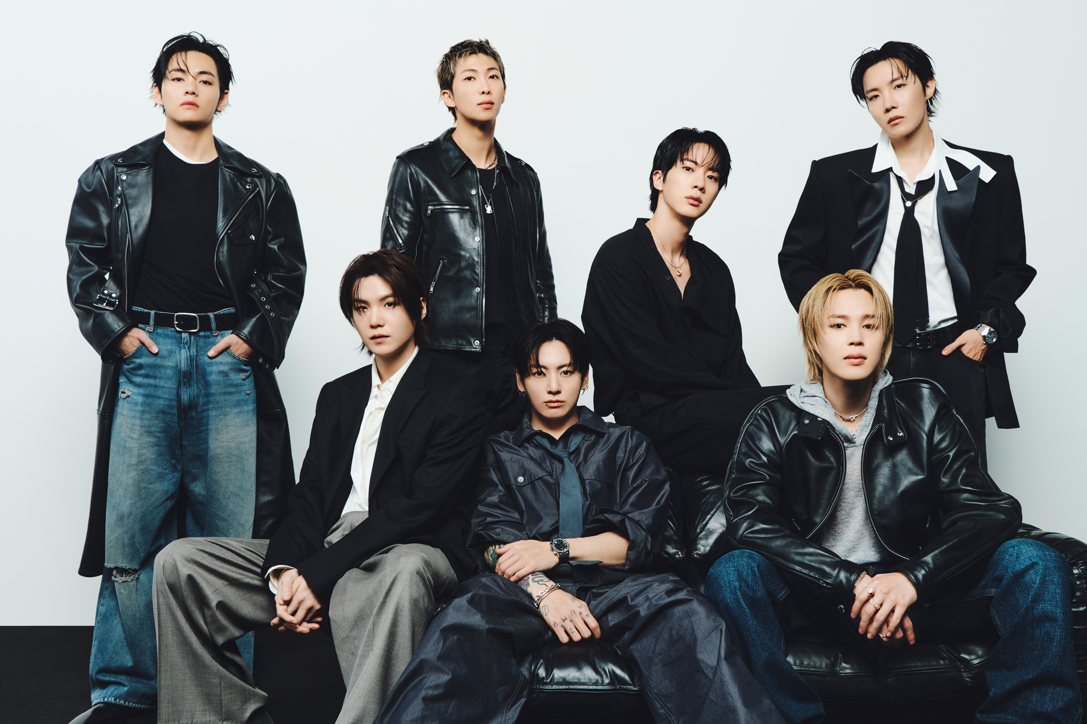 Imagem do BTS, fenômeno mundial. Estão vestindo tons escuros e jeans, três membros sentados e quatro em pé atrás.