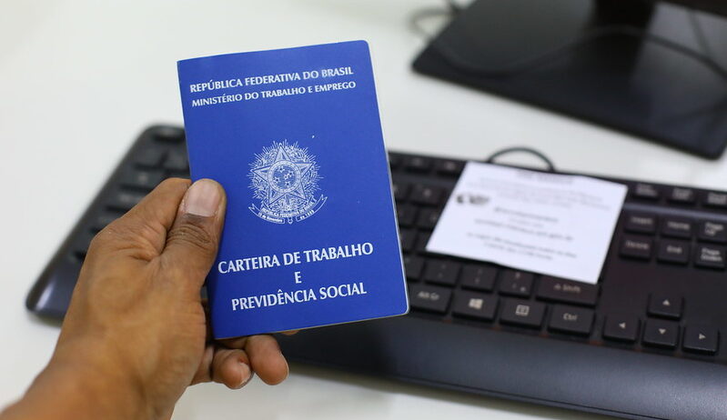 Imagem de uma carteira de trbalho, sendo segurada pela mão de um homem. Ao fundo o teclado de um computador.