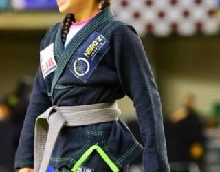 Imagem de uma jovem usando kimono preto e faixa cinza, está no tatame.