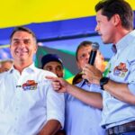 Imagem de Fávio Bolsonaro e Daniel Vilela a frente em um palanque político, Daniel está com microfone em uma das mãos e Flávio está olhando para o público e sorrindo, ao fundo apoiadores.