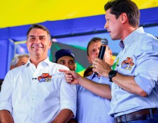 Imagem de Fávio Bolsonaro e Daniel Vilela a frente em um palanque político, Daniel está com microfone em uma das mãos e Flávio está olhando para o público e sorrindo, ao fundo apoiadores.