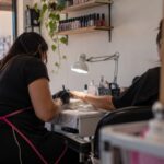Imagem de duas mulheres sentadas, uma delas é manicure e veste um avental, luvas e mascara, a outra está com uma das mãos estendidas, vestindo uma camisa preta. Em cima da mesa, uma luminária, objetos de manicure e ao fundo prateleiras com esmaltes de diversas cores a mostra.