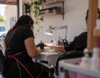 Imagem de duas mulheres sentadas, uma delas é manicure e veste um avental, luvas e mascara, a outra está com uma das mãos estendidas, vestindo uma camisa preta. Em cima da mesa, uma luminária, objetos de manicure e ao fundo prateleiras com esmaltes de diversas cores a mostra.