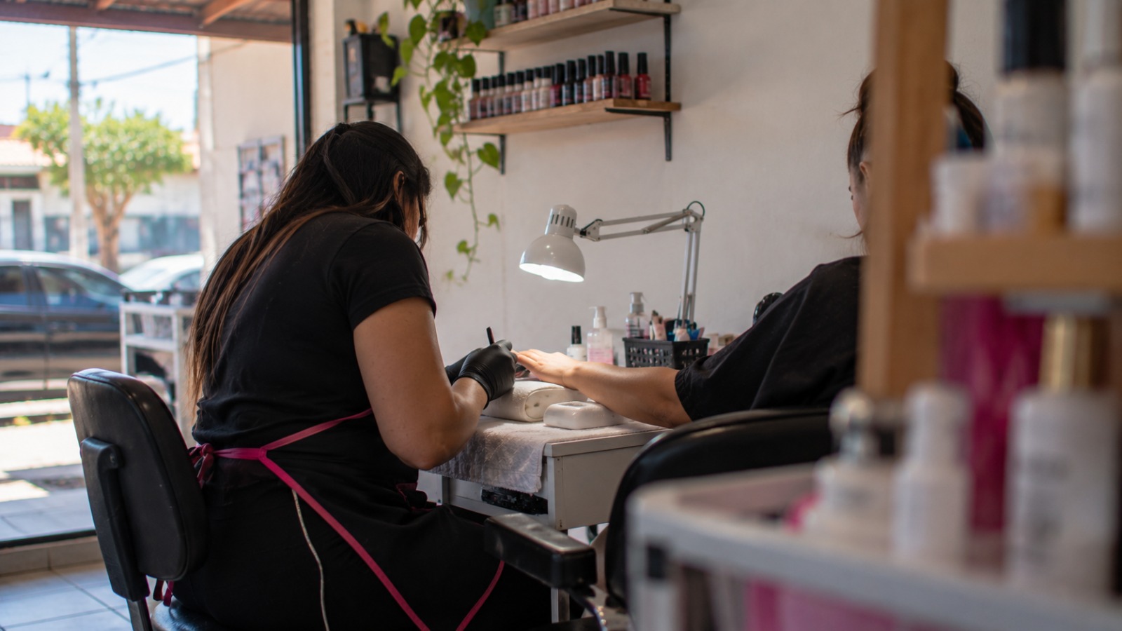 Imagem de duas mulheres sentadas, uma delas é manicure e veste um avental, luvas e mascara, a outra está com uma das mãos estendidas, vestindo uma camisa preta. Em cima da mesa, uma luminária, objetos de manicure e ao fundo prateleiras com esmaltes de diversas cores a mostra.