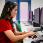 Imagem mulher sentada em frente o computador, ela está de blusa vermelha