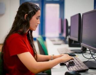 Imagem mulher sentada em frente o computador, ela está de blusa vermelha