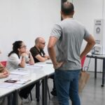 Imagem de local de votação, uma sala de aula, com três mesários e um homem em pé, esperando a sua vez.
