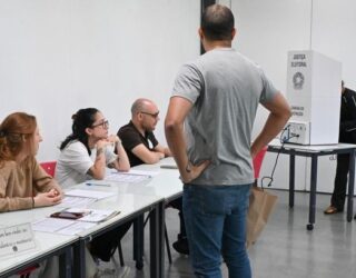 Imagem de local de votação, uma sala de aula, com três mesários e um homem em pé, esperando a sua vez.