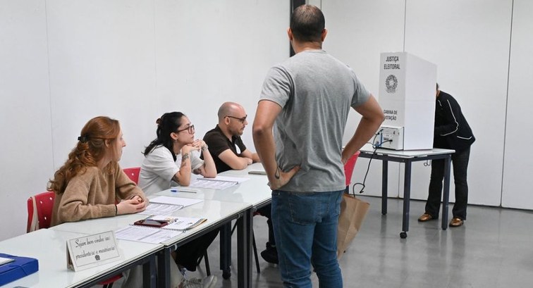 Imagem de local de votação, uma sala de aula, com três mesários e um homem em pé, esperando a sua vez.