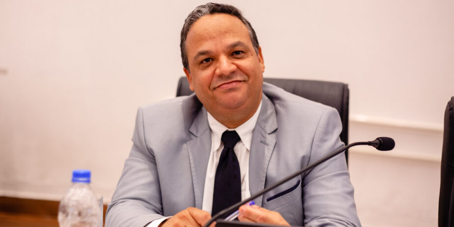 Imagem do vereador Nilson, sentado, olhando para a foto, usanaod terno cinza e gravata preta.