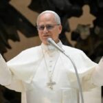 Imagem de Papa Leão XIV de túnica branca, braços abertos e falando no microfone.