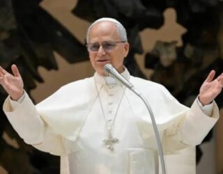 Imagem de Papa Leão XIV de túnica branca, braços abertos e falando no microfone.