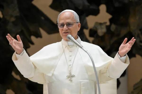 Imagem de Papa Leão XIV de túnica branca, braços abertos e falando no microfone.