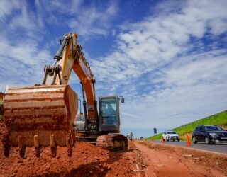 Imagem de uma escavadeira em uma obra, bastante terra vermelha, alguns cones separando obra e asfalto onde há alguns carros.