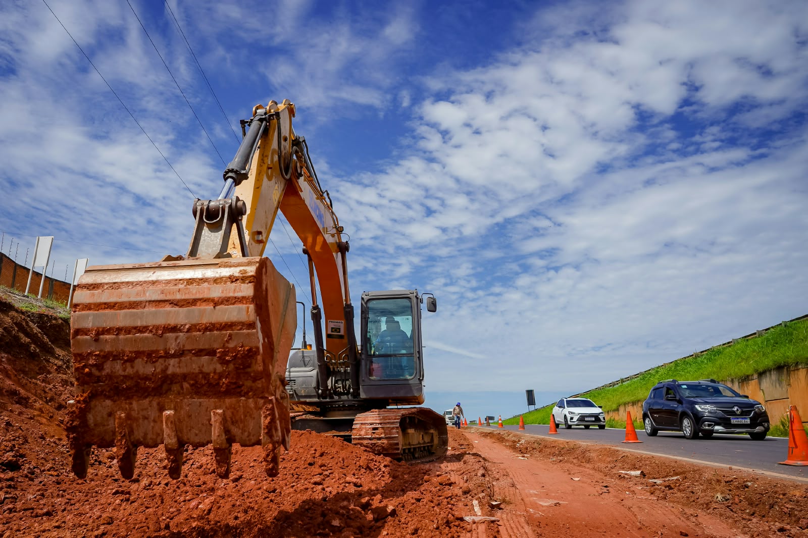 Imagem de uma escavadeira em uma obra, bastante terra vermelha, alguns cones separando obra e asfalto onde há alguns carros.