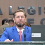 Imagem de um homem branco, olhos claros e de terno azul, falando no microfone. Atrás um letreiro que diz: Assembleia Legislativa