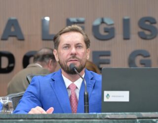 Imagem de um homem branco, olhos claros e de terno azul, falando no microfone. Atrás um letreiro que diz: Assembleia Legislativa