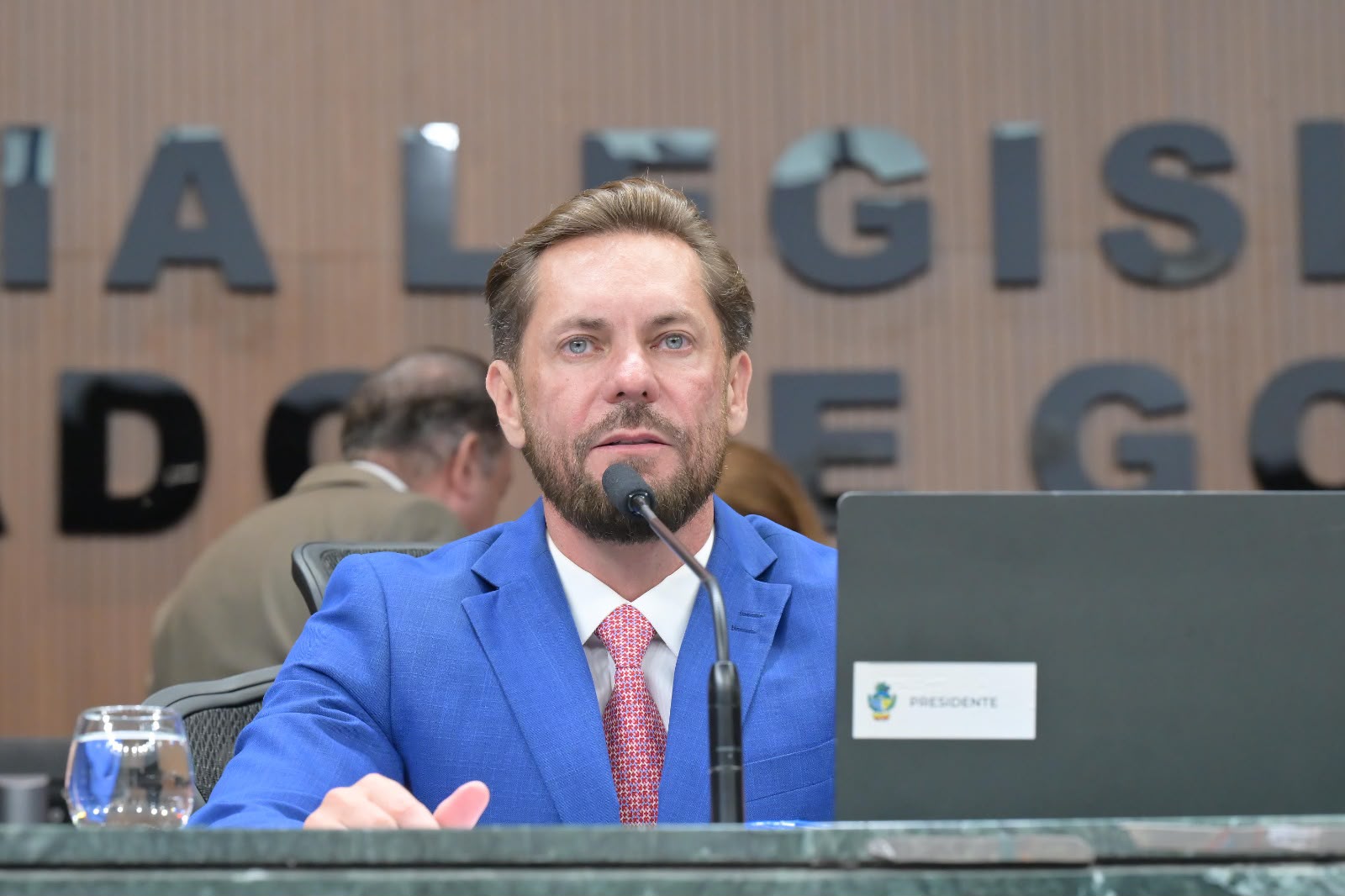 Imagem de um homem branco, olhos claros e de terno azul, falando no microfone. Atrás um letreiro que diz: Assembleia Legislativa