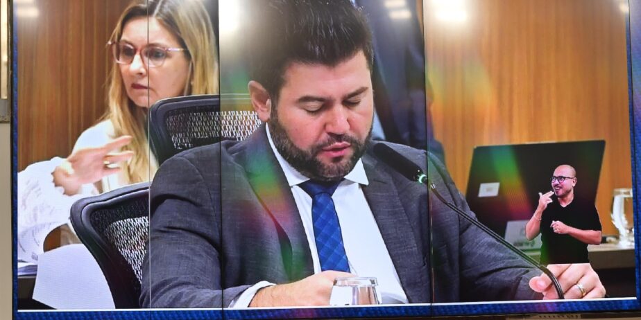Imagem de um homem em um telão de terno cinza, gravata azul e camisa branca, olhando para baixo e falando em um microfone.