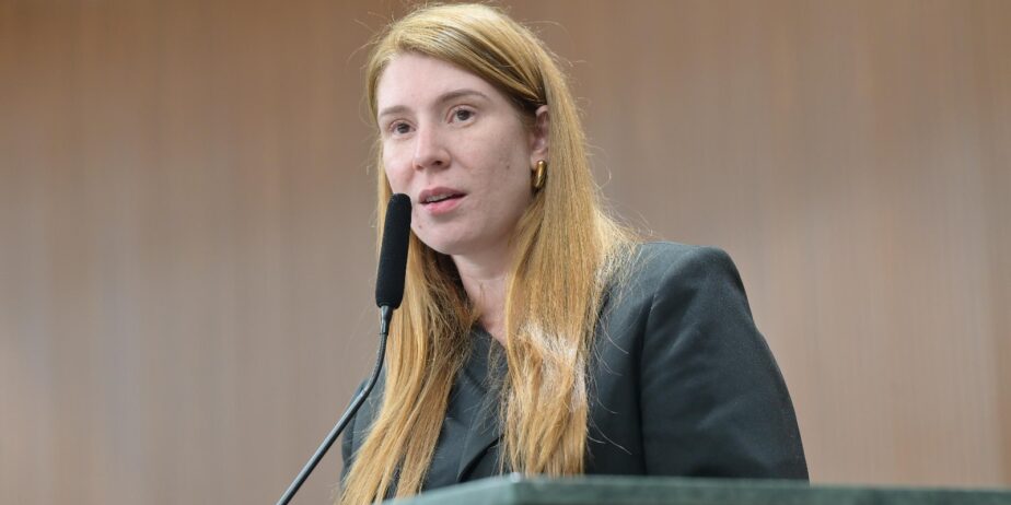 Imagem de uma mulher ruiva, cabelos longos e blazer preto, falando no microfone.