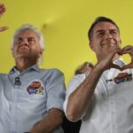 Imagem Caiado e Flávio Bolsonaro em um evento: o da esquerda, de camisa azul e cabelo branco, acena com a mão erguida, enquanto o da direita, de camisa branca, faz o gesto de um coração com as mãos.