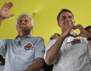 Imagem Caiado e Flávio Bolsonaro em um evento: o da esquerda, de camisa azul e cabelo branco, acena com a mão erguida, enquanto o da direita, de camisa branca, faz o gesto de um coração com as mãos.
