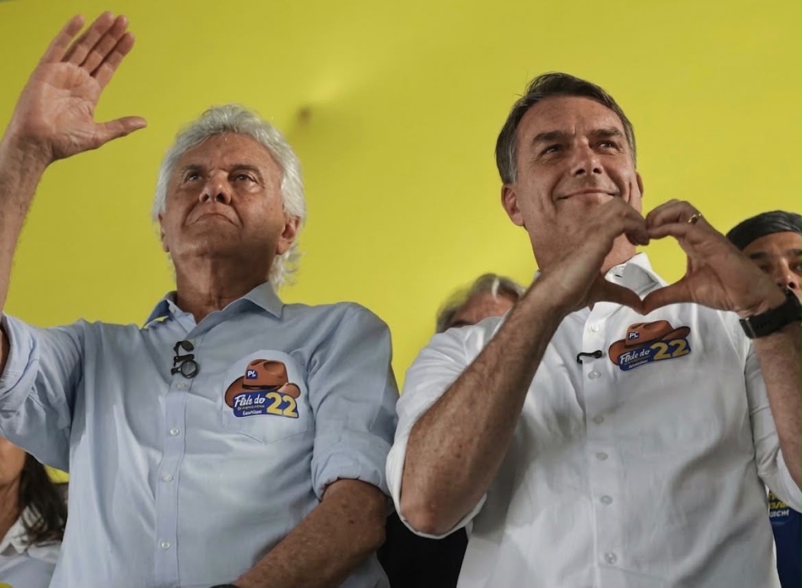 Imagem Caiado e Flávio Bolsonaro em um evento: o da esquerda, de camisa azul e cabelo branco, acena com a mão erguida, enquanto o da direita, de camisa branca, faz o gesto de um coração com as mãos.