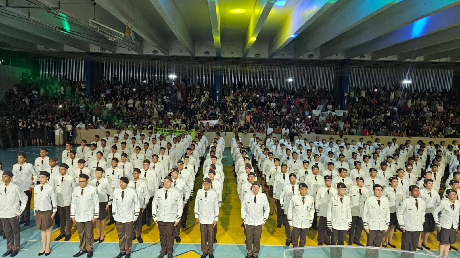 Imagem de alunos de Colégio Militar em posição de sentido em uma quadra da escola, vestindo farda branca.