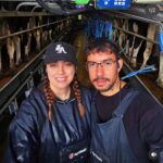 Imagem de um casal, a moça usa tranças no cabelo, boné, roupa preta e avental. O homem está de óculos de grau, roupa preta e avental. Ao fundo o local de trabalho com inúmeras vacas leiteiras.