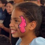 Imagem, criança sorridente com uma borboleta rosa pintada em um lado do rosto.