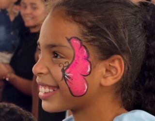 Imagem, criança sorridente com uma borboleta rosa pintada em um lado do rosto.