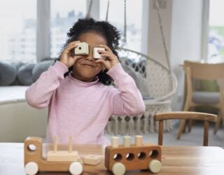 Imagem de uma criança brincando com peças, nas duas mãos, cada uma tem uma pecinha e ela coloca na altura dos olhos, em cima da mesa, brinquedos e atrás dela um balanço.